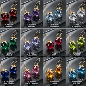May include: A collection of twelve pairs of faceted spherical gemstone earrings with gold-tone hooks and caps, labeled by month and stone: 1. January Garnet, 2. February Amethyst, 3. March Aquamarine, 4. April Crystal Quartz, 5. May Emerald, 6. June Alexandrite, 7. July Ruby, 8. August Peridot, 9. September Blue Sapphire, 10. October Pink Tourmaline, 11. November Citrine, 12. December Blue Topaz.