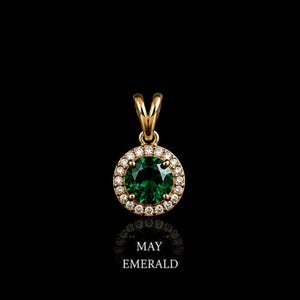 May include: A gold pendant featuring a round, faceted green gemstone at the center, surrounded by a halo of small, clear white stones. The pendant has a gold bail at the top. Below the pendant, the text reads MAY EMERALD in white capital letters.