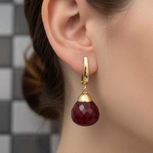May include: A gold-coloured earring featuring a teardrop-shaped, faceted, dark red gemstone. The earring hangs from a gold-coloured hook. The gemstone is capped with gold-coloured metal.