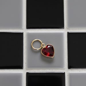 May include: A gold heart-shaped charm with a deep red gemstone. The charm hangs from a small gold ring. The background is a grid of black, grey, and white square tiles.
