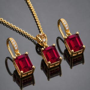 Op de afbeelding: Een gouden sieradenset bestaande uit een ketting met hanger en twee bijpassende oorbellen. Elk sieraad bevat een rechthoekige, smaragdgeslepen dieprode edelsteen in een gouden zetting, met kleine heldere accentstenen op de oorhaken.