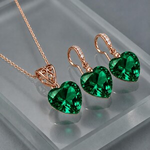 May include: A rose gold jewellery set consisting of a necklace and two earrings. Each piece features a faceted heart-shaped emerald green gemstone. The necklace pendant and earring hooks are accented with small clear stones in a rose gold setting.