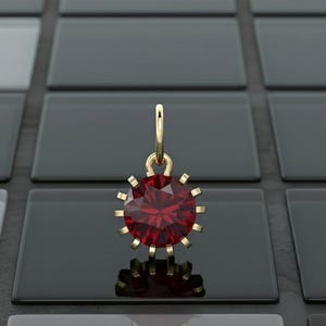 May include: A gold pendant featuring a large, faceted, round, deep red gemstone. The pendant has a sunburst design with gold prongs around the gem. The pendant is suspended from a gold loop.