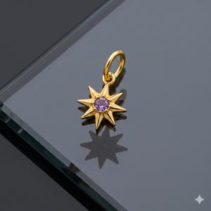 May include: A gold-colored starburst charm with a purple gemstone center. The charm has a loop for attaching to a necklace or bracelet. The starburst design features pointed rays radiating from the center, creating a celestial aesthetic.