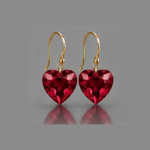 May include: A pair of drop earrings featuring faceted, heart-shaped red gemstones suspended from gold-toned curved ear wires. The stones exhibit a deep crimson hue with multiple reflective facets that catch the light, creating a symmetrical design.