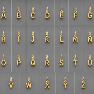 Op de afbeelding: Een collectie van zesentwintig individuele letterbedels van goudkleurig metaal, gerangschikt in alfabetische volgorde van A tot Z. Elke bedel heeft een gepolijst, afgerond oppervlak en een klein ringetje aan de bovenkant voor bevestiging.