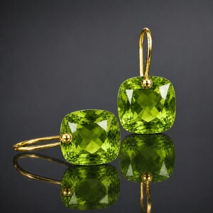 May include: A pair of drop earrings featuring square-cut, faceted lime green gemstones set in gold metal. Each earring has a curved gold wire hook attached to the top of the stone, showcasing a bright, translucent color and geometric cut.