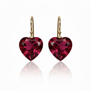 Puede incluir: Un par de pendientes en forma de corazón con una gema de color rojo intenso. Los pendientes tienen ganchos dorados y un diseño facetado. Se muestran sobre un fondo blanco con reflejo.