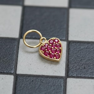 May include: A gold heart pendant, suitable for a necklace. The heart is filled with small, round, red gemstones. The pendant has a loop for a chain and is set against a black and white chequered background.