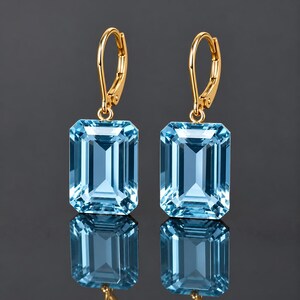 May include: A pair of gold leverback earrings featuring large, rectangular, emerald-cut light blue gemstones. The stones are clear and faceted, suspended from polished gold hooks that secure with a hinged mechanism at the top.