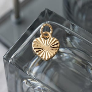 May include: A gold heart-shaped pendant with a textured, sunburst design. The pendant has a twisted loop for attaching to a chain. The charm is displayed on a clear, square glass surface.