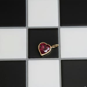 May include: A heart-shaped pendant with a vibrant red gemstone set in a gold-colored bezel. The pendant is displayed against a backdrop of black and white square tiles, creating a striking contrast. The pendant's design is simple yet elegant.