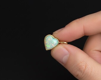 Puede incluir: Un colgante en forma de corazón con un marco dorado. El colgante presenta una piedra de ópalo blanco con motas verdes y azules. La joya se sostiene en la mano de una persona sobre un fondo negro. El colgante es una pequeña joya.