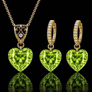 May include: A jewelry set consisting of a necklace and two hoop earrings. Each piece features a faceted heart-shaped lime green gemstone. The necklace pendant has a gold filigree bail with small clear stones, suspended from a gold chain.