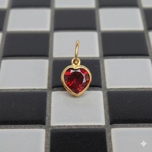 May include: A gold heart pendant with a red gemstone. The heart-shaped gem is set in a gold frame and hangs from a small loop. The background features a checkered pattern of black, white, and grey tiles. The pendant is suitable for a necklace.