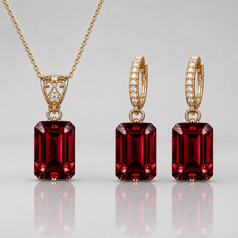 May include: A gold jewelry set consisting of a necklace and a pair of hoop earrings. Each piece features a large, rectangular, deep red gemstone pendant suspended from a gold setting accented with small, clear, sparkling white stones.