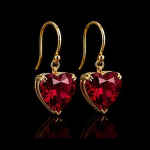 Puede incluir: Un par de pendientes colgantes de oro con piedras preciosas rojas facetadas en forma de corazón. Cada pendiente cuelga de un gancho dorado curvado y se sujeta mediante pequeñas garras de oro que aseguran la piedra roja en la base.