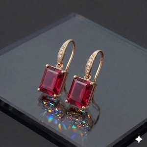 Può includere: Un paio di orecchini pendenti color oro con pietre preziose rettangolari rosso intenso. Gli orecchini hanno un design delicato con piccoli accenti scintillanti lungo la parte superiore curva. Gli orecchini sono esposti su una superficie riflettente.