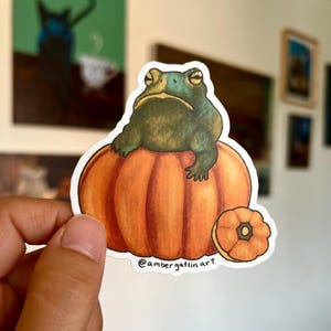 May include: A sticker featuring a green frog sitting on an orange pumpkin. A smaller pumpkin is next to the larger one. The sticker has a white border and the text "@ambergatlinart" is printed on the bottom.