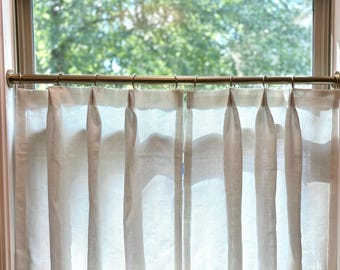 May include: Sheer, off-white linen curtains hanging from a brass rod in front of a window. The curtains have a simple design with a neutral-colored border at the bottom. The window looks out onto a lush green tree.