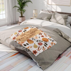 Può includere: Una coperta decorativa con un design a tema autunnale con zucche, pere ed elementi floreali. La coperta ha la scritta "ARE YOU FALL-O-WEEN JESUS?". La coperta è appoggiata su un letto con biancheria da letto dai colori neutri.
