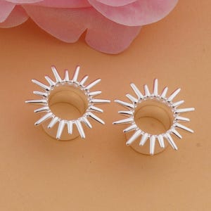 May include: Two silver-colored ear gauges with a sunburst design. Each gauge has a circular center with radiating spikes. The gauges are displayed on a light orange surface, with a pink flower in the background.