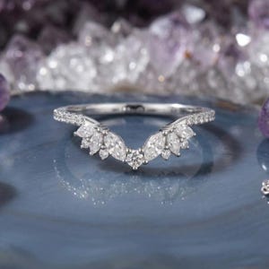 Op de afbeelding: Een zilverkleurige, gebogen ring met een reeks heldere stenen in marquise- en ronde slijpvorm, gerangschikt in een V-vormig cluster aan de bovenkant. De ringband is dun en bezet met kleine heldere steentjes langs het zichtbare bovendeel.