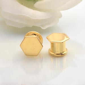 Puede incluir: Dos tapones para los oídos hexagonales dorados. Los tapones tienen un diseño geométrico plano y un acabado brillante. Los tapones se muestran sobre una superficie blanca con una flor blanca borrosa en el fondo.