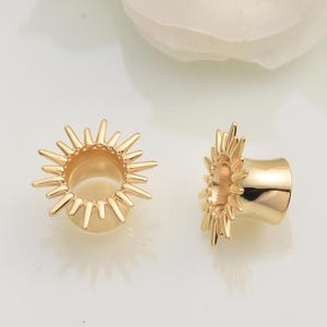 Op de afbeelding: Twee goudkleurige oortunnels met een sunburst-ontwerp. De tunnels zijn cilindrisch met een uitlopende rand en een reeks puntige stralen die naar buiten uitsteken. De sieraden worden op een witte achtergrond getoond.