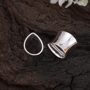 May include: Two silver-colored ear gauges. One is a teardrop shape, and the other is a flared cylinder. The jewelry is highly reflective, with a polished finish. The gauges are displayed on a dark, textured surface.
