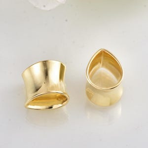 May include: Two gold-colored, open-ended ear tunnels. One is a curved, cylindrical shape, and the other is a teardrop shape. Both have a polished, reflective surface, and are set against a white background.