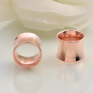 Könnte beinhalten: Zwei roségoldfarbene Tunnel-Ohrstecker. Einer ist auf beiden Seiten abgerundet, der andere hat eine ausgestellte, sanduhrförmige Form. Die Dehnungsstecker sind glatt und reflektierend, mit poliertem Finish. Der Hintergrund ist ein weiches, verschwommenes Weiß.