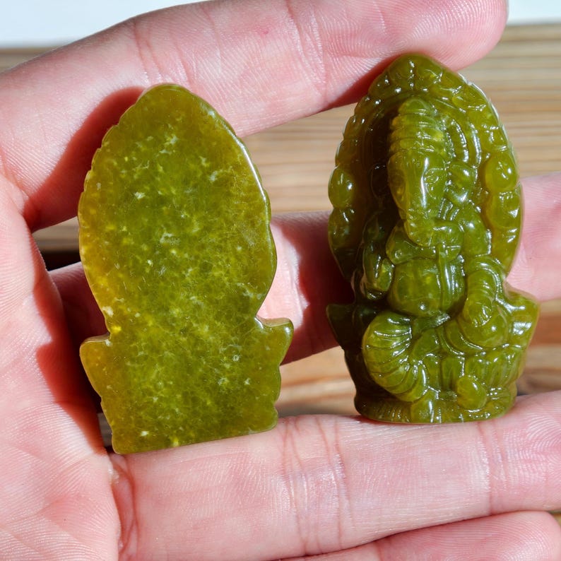 Puede incluir: Se muestran dos tallas de jade verde. Una es una forma plana y abstracta, y la otra es una escultura detallada de una deidad con cabeza de elefante. Las tallas se sostienen en una mano, mostrando su tama&ntilde;o y color.