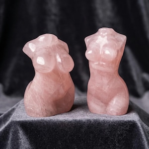 Puede incluir: Dos esculturas de cuarzo rosa del torso femenino. Las esculturas son de color rosa suave y tienen superficies lisas y pulidas. Los bustos se exhiben sobre una superficie de terciopelo gris oscuro.