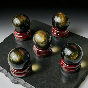 Könnte beinhalten: Fünf goldene Obsidiankugeln auf roten Holzständern, angeordnet auf einer dunkelgrauen Oberfläche. Die Kugeln haben eine reflektierende, polierte Oberfläche mit goldenen und schwarzen Wirbeln. Die Ständer sind rund und glänzend.