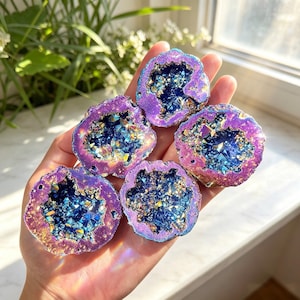 May include: Five small, circular geode slices with iridescent purple and gold outer edges. The centers are filled with deep blue, crystalline structures that reflect light, creating a sparkling, multi-faceted appearance across the cluster.
