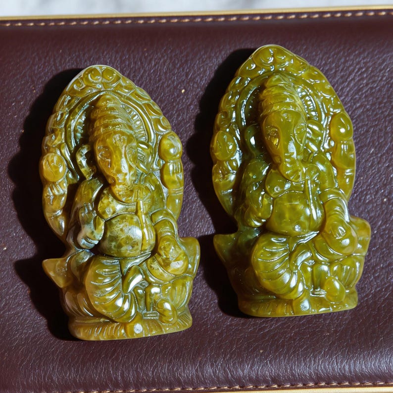 Puede incluir: Dos esculturas de color jade del dios hind&uacute; Ganesha. Cada escultura representa a Ganesha sentado, con cabeza de elefante y m&uacute;ltiples brazos, rodeado de un tel&oacute;n de fondo arqueado y ornamentado. Las esculturas est&aacute;n sobre una superficie marr&oacute;n.