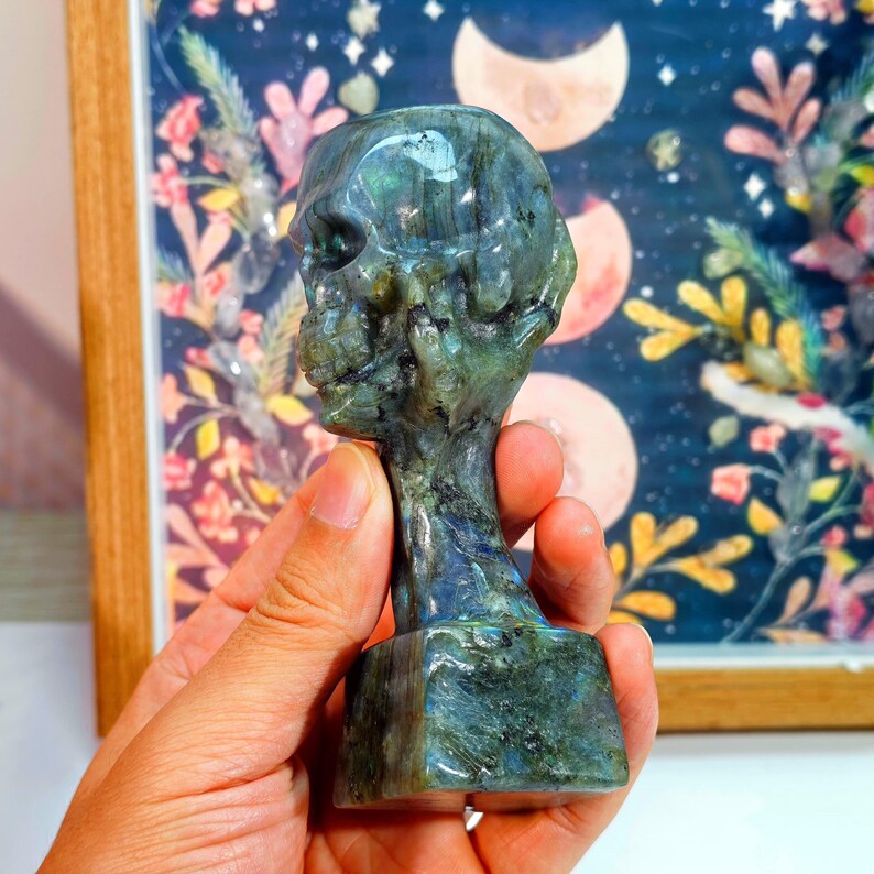Puede incluir: Una escultura de calavera de labradorita tallada. La calavera es una mezcla de tonos azules, verdes y grises. Se sostiene en una mano, con un fondo decorativo con lunas y elementos florales. La escultura tiene una base rectangular.