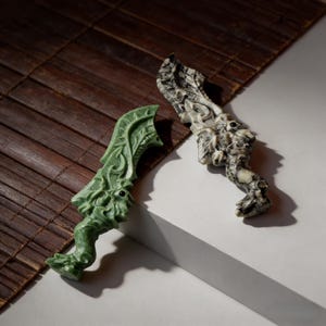 May include: Two carved stone daggers. One is green, the other is black and white. Both daggers have a dragon-like design with intricate details. The daggers are placed on a white surface with a brown bamboo mat in the background.