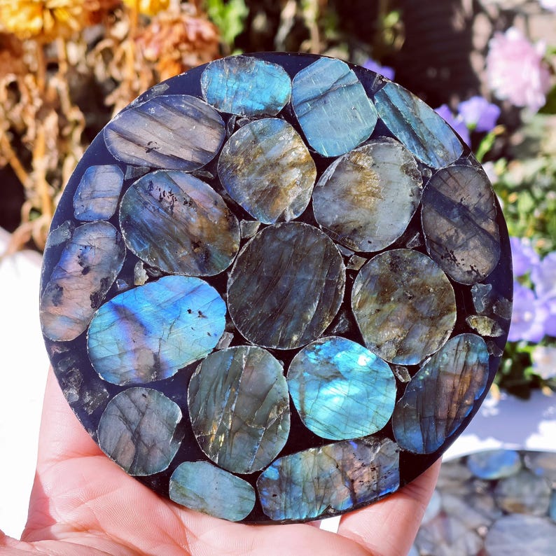 May include: Round coaster with a mosaic design. The coaster is made of dark resin and inlaid with polished labradorite stones in shades of blue, gray, and gold. The stones are arranged in a circular pattern.