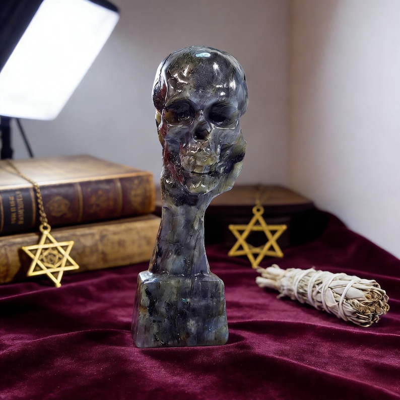 Puede incluir: Una escultura de calavera tallada en tonos grises y verdes, montada sobre una base rectangular. La escultura se exhibe sobre una superficie de terciopelo burdeos, con un colgante dorado de la Estrella de David y un manojo de salvia cerca.