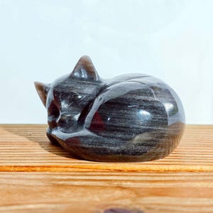 May include: A polished, dark gray cat figurine in a curled-up sleeping position. The stone cat has a smooth, reflective surface with subtle streaks of lighter gray. The figurine is resting on a wooden surface.