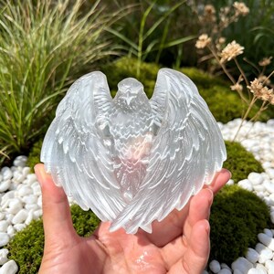 May include: A translucent, white, carved crystal sculpture in the shape of an eagle with its wings spread wide. The carving features detailed feather textures across the wings and body, creating a clear, reflective surface with a smooth finish.