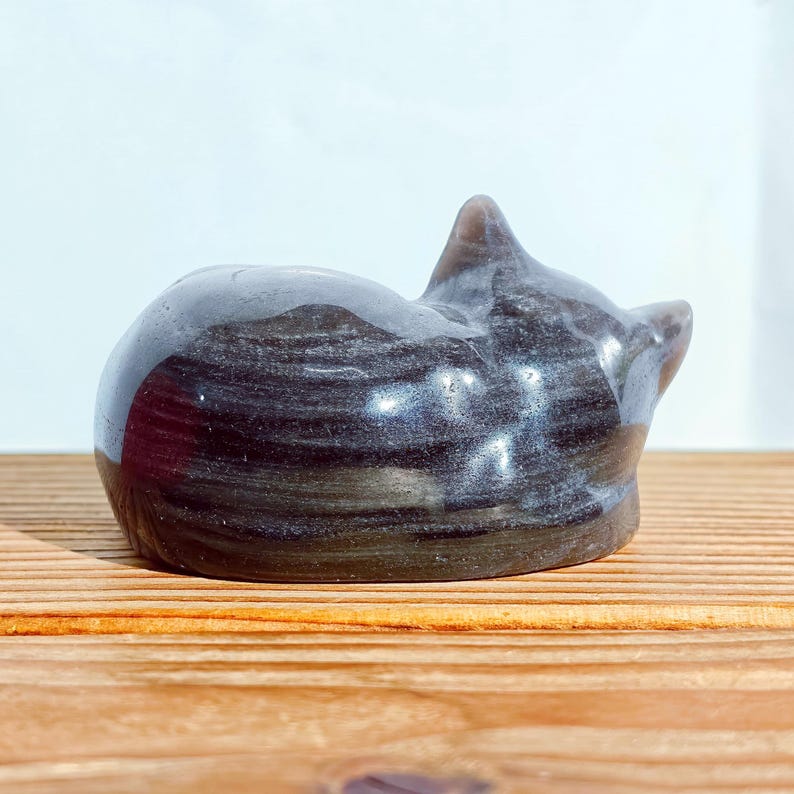 May include: A dark gray cat figurine in a curled-up position. The smooth, polished surface of the cat has subtle streaks of lighter gray. The figurine is resting on a wooden surface, with a plain white background.