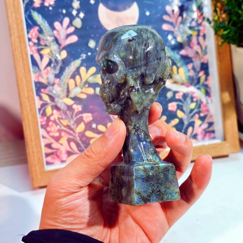 Puede incluir: Una figura de calavera tallada en labradorita iridiscente. La calavera tiene detalles y se asienta sobre una base rectangular. La piedra muestra tonos de azul, verde y gris, con un acabado pulido. La figura se sostiene en una mano.
