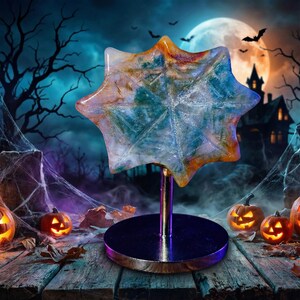 May include: A decorative star-shaped ornament with a marbled effect in blue, orange, and white, mounted on a silver stand with a circular base. The background depicts a Halloween scene with pumpkins, a full moon, and a haunted house.