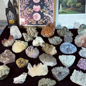 Peut inclure: Une collection de cristaux et de pierres précieuses de différentes formes et couleurs, dont du quartz clair, de l'améthyste et de la pyrite. Les cristaux sont disposés sur une surface noire, avec une impression encadrée d'un motif céleste et une maison décorative d'Halloween en arrière-plan.