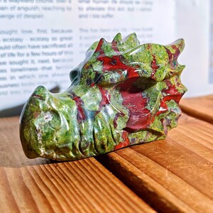 May include: A carved stone dragon head sculpture. The dragon head is green with red veining and details. The sculpture is resting on a wooden surface. The dragon head is a unique decorative item.