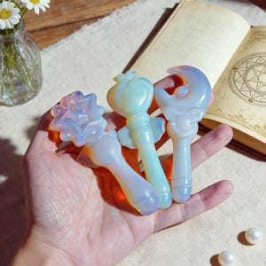 Può includere: Tre bacchette di cristallo opalescente. Le bacchette, tenute in una mano, presentano un design a stella, un design a forma di cuore e una luna crescente. Le bacchette sono bianche, blu e gialle.