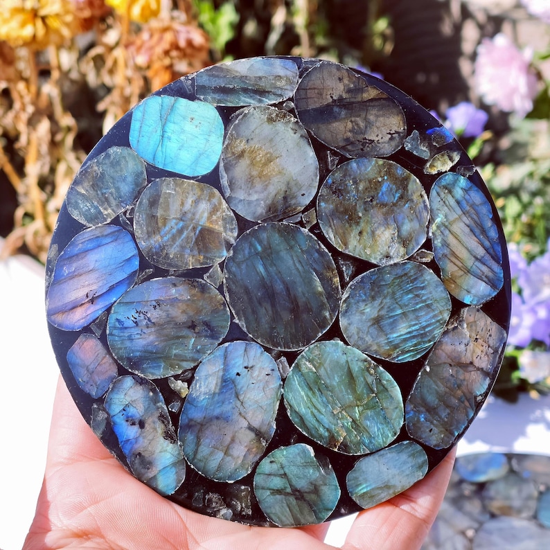 May include: A round, polished stone coaster with a mosaic of iridescent labradorite stones in shades of blue, teal, and gray. The stones are set in a dark, possibly black, resin base. The coaster is approximately 4 inches in diameter.